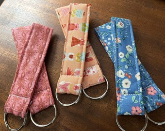 May include: Three fabric keychains with different patterns. The keychain on the left has a pink and red geometric pattern. The keychain in the middle has a peach background with a pattern of colorful dresses. The keychain on the right has a blue background with a white floral pattern.