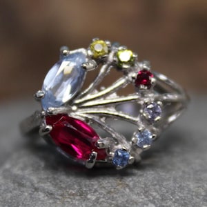 May include: A silver ring with a delicate, branching design set with colorful gemstones. The ring features a large, oval-shaped blue stone, two smaller red stones, and several smaller yellow and white stones.