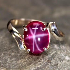May include: A silver ring featuring a polished, oval-shaped, deep magenta star gemstone. The stone displays a distinct six-rayed star pattern across its surface. The metal band has a smooth, curved design that tapers as it meets the stone setting.