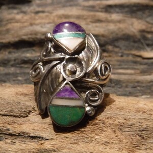 May include: A silver ring with a swirling design and three stones. The stones are a teardrop shape and are purple, green, and white.