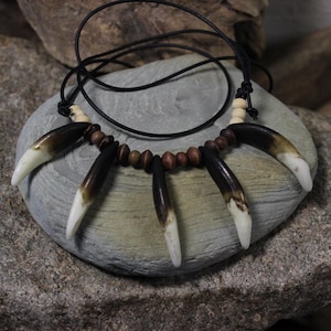 May include: A handcrafted necklace featuring a series of dark brown and white teeth-shaped pendants. The pendants are strung on a black cord, interspersed with small wooden beads. The necklace is displayed on a gray stone surface.