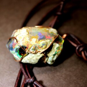 May include: A rough opal gemstone pendant, secured with copper wire and strung on a brown leather cord. The opal exhibits iridescent colours, including green, blue, and red. A unique, handcrafted piece of jewellery.