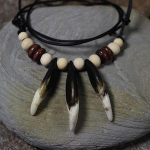 May include: A handcrafted necklace featuring three dark, pointed pendants with white tips, interspersed with cream-colored and dark brown wooden beads. The necklace is strung on a black cord, creating a rustic and natural aesthetic.