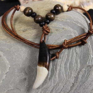May include: A brown leather cord necklace with a pendant shaped like a tooth. The tooth pendant is two-toned, with a white tip and a dark brown top. The necklace also features wooden beads in various shades of brown and black.