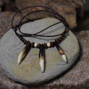May include: A handcrafted necklace featuring three large, pointed pendants with a gradient of brown to white. The necklace includes small, dark brown wooden beads and a brown leather cord. The necklace is displayed on a gray stone.