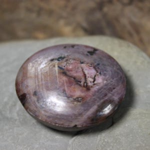 May include: A polished, round gemstone with a blend of purple, pink, and brown hues. The stone has a smooth, glossy surface with natural patterns and variations in color. The gemstone is set against a neutral background.