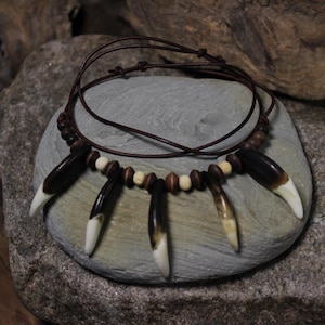 May include: A handcrafted necklace featuring dark brown and cream-colored teeth-shaped pendants. The pendants are strung on a brown leather cord, interspersed with wooden beads. The necklace is displayed on a gray stone surface.