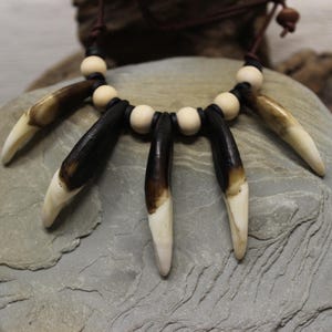 May include: A necklace featuring a series of dark brown and cream-colored pendants, resembling teeth or claws, strung together with black cord and wooden beads. The necklace is displayed on a gray stone surface.