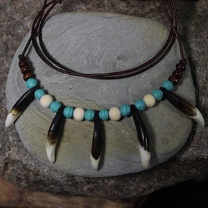 May include: A handcrafted necklace featuring a brown leather cord. The necklace is adorned with alternating turquoise and cream-colored beads, dark brown beads, and dark brown pendants with white tips. The necklace is displayed on a gray stone.