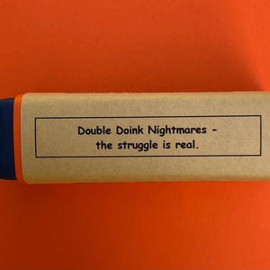 以下が含まれることがあります： 青い縁取りのある茶色の段ボール箱。箱には「Double Doink Nightmares - the struggle is real.」と書かれたラベルが付いています。
