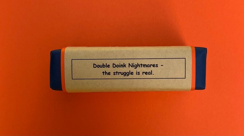 以下が含まれることがあります： 青い縁取りのある茶色の段ボール箱。箱には「Double Doink Nightmares - the struggle is real.」と書かれたラベルが付いています。