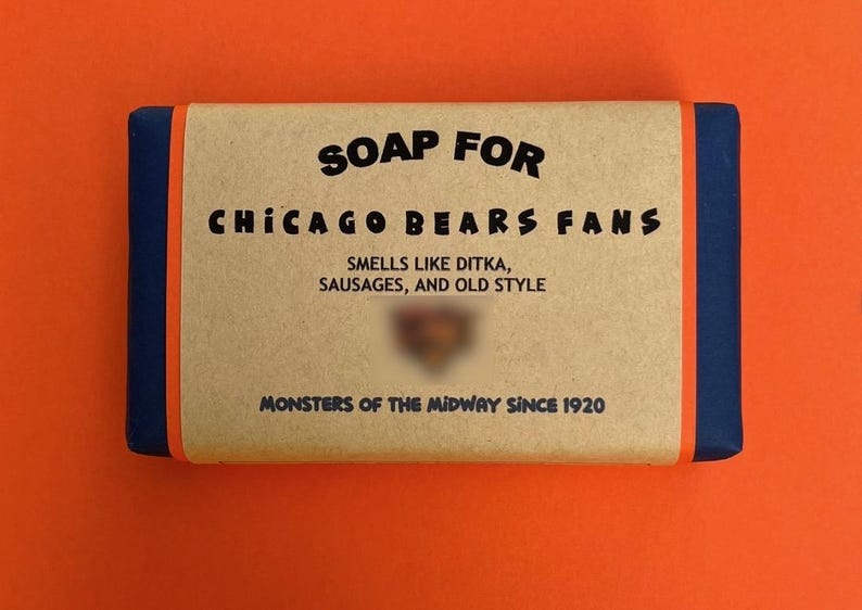 以下が含まれることがあります： 青い縁取りのある茶色の石鹸。石鹸には「SOAP FOR CHICAGO BEARS FANS」と「SMELLS LIKE DITKA, SAUSAGES, AND OLD STYLE」のラベルが貼られています。ラベルの下部には「MONSTERS OF THE MIDWAY SINCE 1920」と書いてあります。