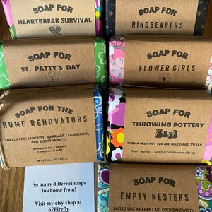 May include: A collection of nine handmade soap bars with various scents and themes. The soaps are wrapped in colorful patterned paper and have brown paper labels with white text. The labels include the names of the soaps, such as "Soap for Heartbreak Survival", "Soap for Ring Bearers", and "Soap for Empty Nesters".