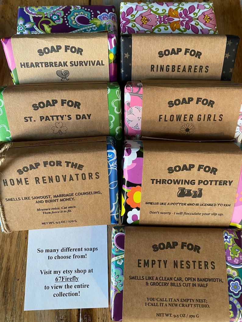 May include: A collection of nine handmade soap bars with various scents and themes. The soaps are wrapped in colorful patterned paper and have brown paper labels with white text. The labels include the names of the soaps, such as "Soap for Heartbreak Survival", "Soap for Ring Bearers", and "Soap for Empty Nesters".