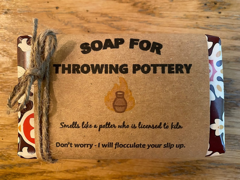 May include: A brown paper wrapped bar of soap with a red and white patterned paper border. The label reads "SOAP FOR THROWING POTTERY" with a small illustration of a clay pot and flames. The text below reads "Smells like a potter who is licensed to kiln. Don't worry - I will flocculate your slip up."