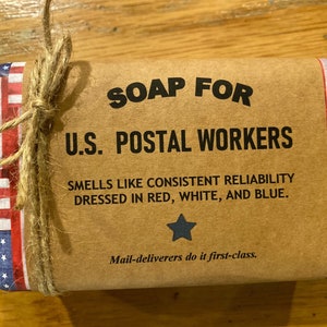 May include: A brown paper wrapped bar of soap with a red, white, and blue patterned border. The label reads "SOAP FOR U.S. POSTAL WORKERS SMELLS LIKE CONSISTENT RELIABILITY DRESSED IN RED, WHITE, AND BLUE. Mail-deliverers do it first-class."