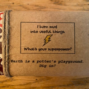 May include: A brown paper package with a colorful patterned design on the sides. The package is tied with twine and has a label that reads "I turn mud into useful things. What's your superpower? Earth is a potter's playground. Dig in!"