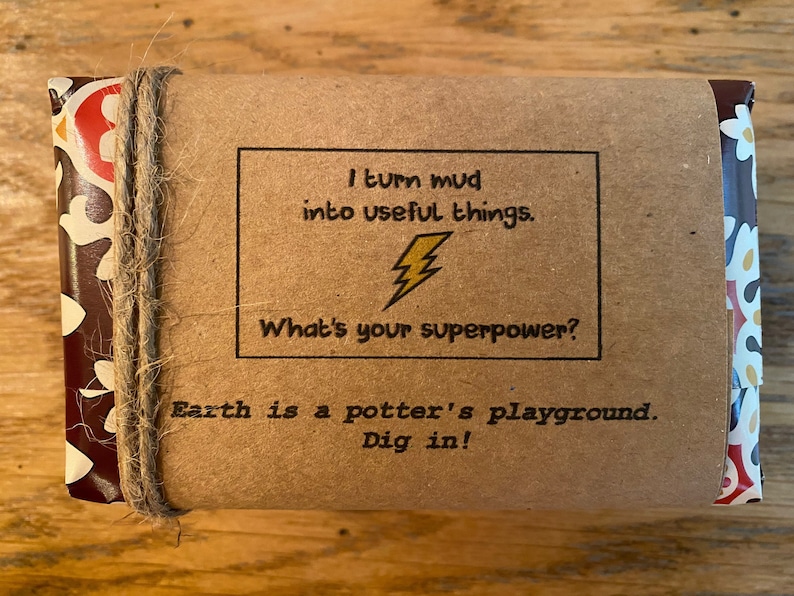 May include: A brown paper package with a colorful patterned design on the sides. The package is tied with twine and has a label that reads "I turn mud into useful things. What's your superpower? Earth is a potter's playground. Dig in!"
