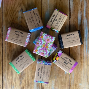 May include: Assortment of eight handmade soap bars wrapped in colorful fabric with various scents and names. The soap bars are arranged on a wooden surface. The soap bar names include "Soap for Flower Girls", "Soap for Home Renovators", "Soap for Heartbreak Survival", "Soap for Ring Bearers", "Soap for St. Patty's Day", "Soap for Empty Nesters", "Soap for Throwing Pottery".