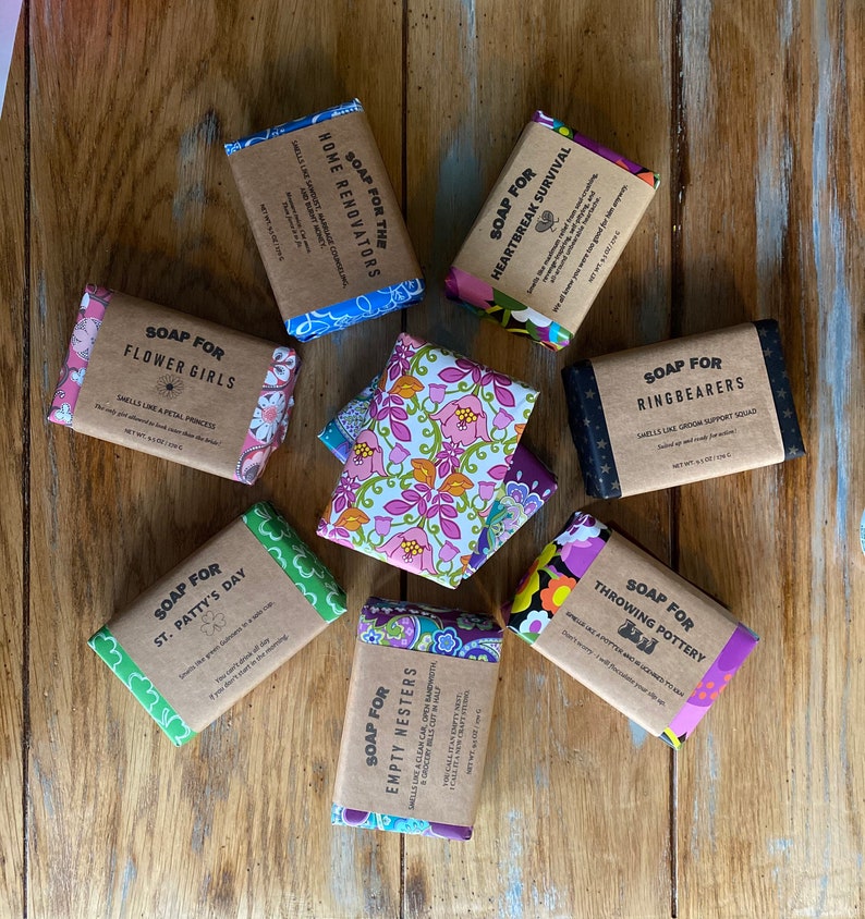 May include: Assortment of eight handmade soap bars wrapped in colorful fabric with various scents and names. The soap bars are arranged on a wooden surface. The soap bar names include "Soap for Flower Girls", "Soap for Home Renovators", "Soap for Heartbreak Survival", "Soap for Ring Bearers", "Soap for St. Patty's Day", "Soap for Empty Nesters", "Soap for Throwing Pottery".