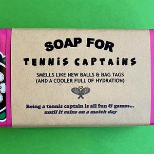 May include: A bar of soap with a brown paper label that reads "SOAP FOR TENNIS CAPTAINS SMELLS LIKE NEW BALLS & BAG TAGS (AND A COOLER FULL OF HYDRATION) Being a tennis captain is all fun & games... until it rains on a match day". The soap is wrapped in pink paper with a black and white floral pattern.