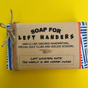 May include: A brown bar of soap with blue and white stripes on the edges. The soap is wrapped in brown paper with the text "SOAP FOR LEFT HANDERS" in black lettering. The text below reads "SMELLS LIKE SMEARED HANDWRITING, SPECIAL GOLF CLUBS AND USELESS SCISSORS."  The bottom of the wrapper has the text "LEFT HANDERS UNITE! THE WORLD IS OUR MIRROR IMAGE" in black lettering.