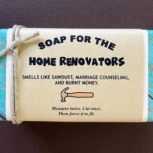 May include: A brown bar of soap with a blue and white patterned border. The soap has the text "SOAP FOR THE HOME RENOVATORS" in black lettering. The text below reads "SMELLS LIKE SAWDUST, MARRIAGE COUNSELING, AND BURNT MONEY."  The bottom of the soap has the text "Measure twice. Cut once. Then force it to fit."