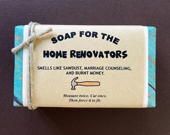 May include: A brown bar of soap with a blue and white patterned border. The soap has the text "SOAP FOR THE HOME RENOVATORS" in black lettering. The text below reads "SMELLS LIKE SAWDUST, MARRIAGE COUNSELING, AND BURNT MONEY."  The bottom of the soap has the text "Measure twice. Cut once. Then force it to fit."