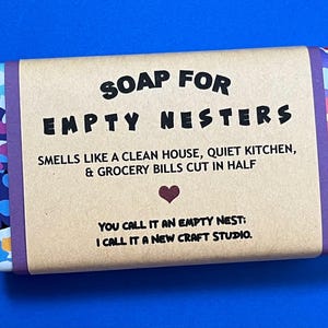 May include: A brown bar of soap with a purple and pink floral design on the edges. The soap is wrapped in a brown paper label with the text "SOAP FOR EMPTY NESTERS" in black. The label also includes the text "SMELLS LIKE A CLEAN HOUSE, QUIET KITCHEN, & GROCERY BILLS CUT IN HALF" and "YOU CALL IT AN EMPTY NEST: I CALL IT A NEW CRAFT STUDIO."