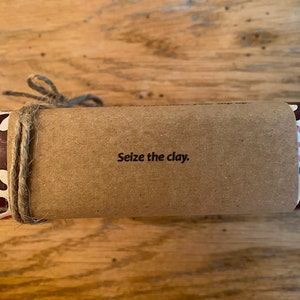 May include: A brown paper-wrapped package with a colorful patterned end. The package is tied with twine and has the text "Seize the clay." printed on the front.