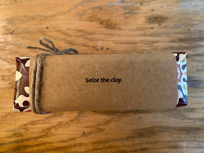 May include: A brown paper-wrapped package with a colorful patterned end. The package is tied with twine and has the text "Seize the clay." printed on the front.