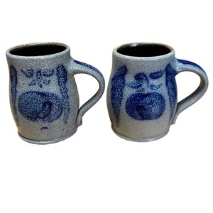 Può includere: Due tazze in gres con finitura grigia screziata e disegni floreali blu scuro. Ogni tazza ha un interno marrone scuro e un manico curvo. Le tazze sono probabilmente fatte a mano e adatte per bevande calde.