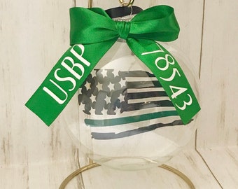 May include: Clear glass Christmas ornament with a painted American flag design. The ornament is adorned with a green ribbon featuring the text "USBP 18543". It hangs from a gold-colored metal stand.