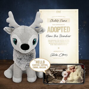 May include: A plush gray reindeer toy with large green eyes and a fluffy chest, next to an adoption certificate. The certificate reads "Adopted Hani the Reindeer" and is approved by Santa Claus. A phone displays a video.