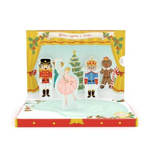 Peut inclure: Une carte de Noël pop-up représentant une scène de ballet avec une ballerine, un Casse-Noisette et d'autres personnages. La carte présente une scène avec les mots "Once upon a time..." et "Press here".
