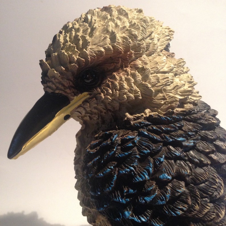 May include: A close-up of a ceramic kookaburra figurine. The bird has a black beak with a yellow stripe, brown and white feathers on the head, and blue and black feathers on the body.