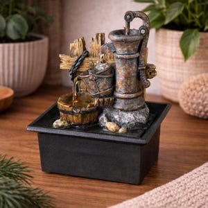 May include: A tabletop water fountain featuring a weathered bronze-coloured hand pump, a small bucket, and a wooden barrel design. The structure sits in a black square basin with water flowing from the pump into the bucket and barrel below.