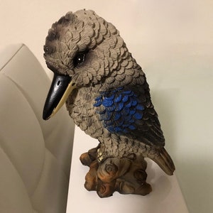 May include: A gray and blue ceramic figurine of a kookaburra bird perched on a tree branch. The bird has a black beak and blue feathers on its wings.