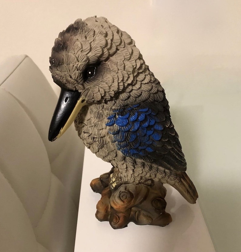 May include: A gray and blue ceramic figurine of a kookaburra bird perched on a tree branch. The bird has a black beak and blue feathers on its wings.
