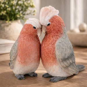 May include: Two bird ornaments in a close embrace. The birds feature a colour palette of red, grey, and white. They are positioned on a wooden surface. The figurines are designed to resemble cockatoos.