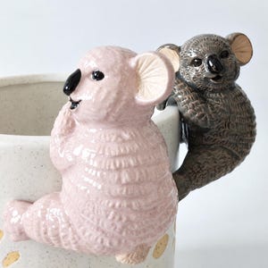 May include: A ceramic mug featuring two sculpted koala figures attached to the rim. One koala is light pink with a black nose, while the other is dark grey with a black nose. The mug itself is off-white with a speckled texture and yellow accents.