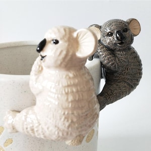May include: A white ceramic mug featuring a koala bear design. The koala is holding a smaller koala bear in its arms. The mug is decorated with brown speckles.
