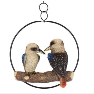 May include: A pair of brown and blue kookaburra birds perched on a branch, surrounded by a black metal ring. The birds are facing each other, with the larger bird on the right and the smaller bird on the left. The ring is suspended from the ceiling by a chain.