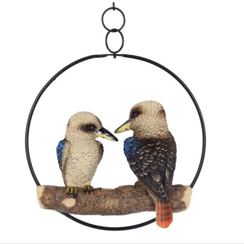 May include: A pair of brown and blue kookaburra birds perched on a branch, surrounded by a black metal ring. The birds are facing each other, with the larger bird on the right and the smaller bird on the left. The ring is suspended from the ceiling by a chain.