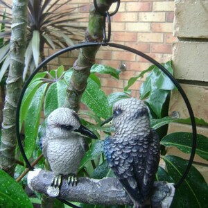 May include: Two gray and blue bird figurines perched on a branch, suspended from a black metal ring. The birds are facing each other, with one looking to the left and the other looking to the right. The branch is surrounded by green leaves.