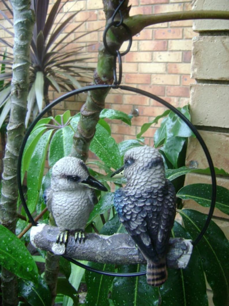 May include: Two gray and blue bird figurines perched on a branch, suspended from a black metal ring. The birds are facing each other, with one looking to the left and the other looking to the right. The branch is surrounded by green leaves.