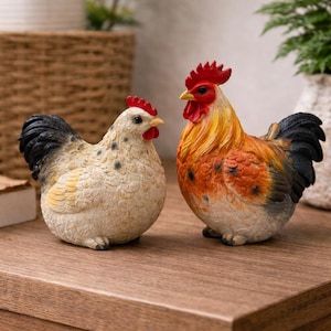 May include: Two ceramic rooster and hen ornaments. The rooster is orange, yellow, and black, while the hen is white with black spots. Both have red combs and beaks. They are displayed on a wooden surface.