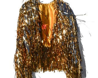 May include: A gold tinsel jacket hangs on a white hanger against a white background. The jacket is covered in long, shimmering gold tinsel strands. The interior lining is a bright yellow-orange color. The jacket is a festive garment.