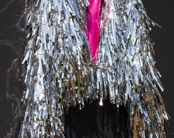 May include: A silver fringe jacket with a bright pink lining hangs against a black background. The jacket is covered in long, shiny silver fringe, creating a textured, eye-catching effect. A small tag with text is visible on the inside.