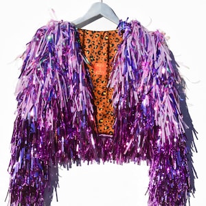 May include: A cropped jacket with a vibrant purple tinsel exterior and a leopard print lining. The jacket is hanging on a white hanger against a white background. The sleeves are long and the jacket is open in the front.